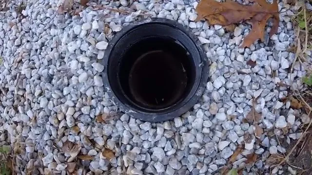 Black cleanout ring with a dark opening set in a bed of gravel with a few fallen leaves