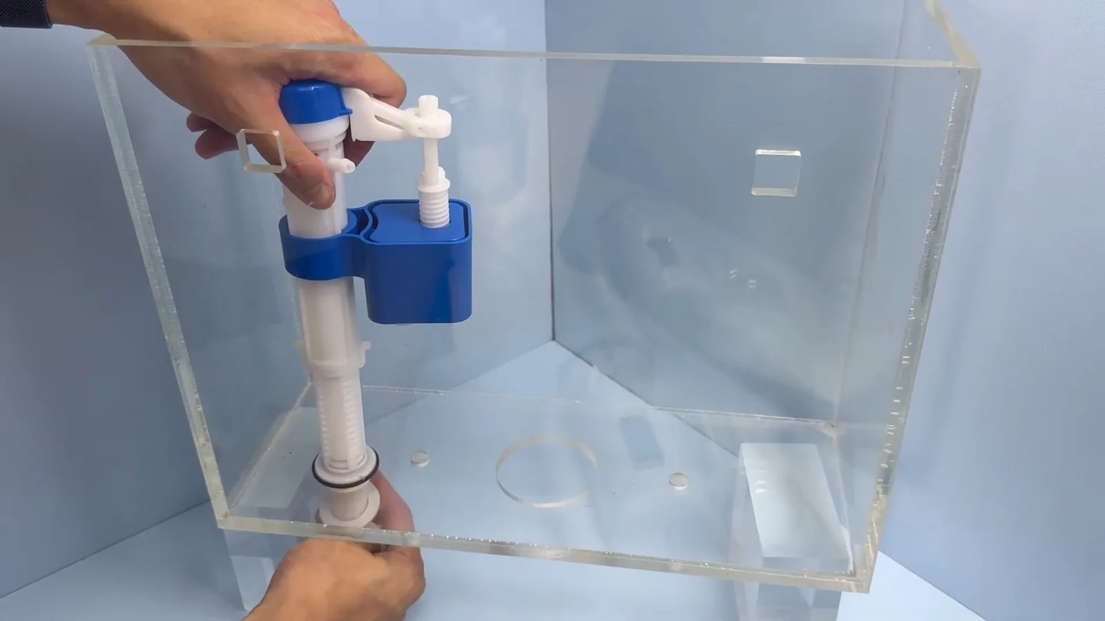 Clear acrylic toilet tank with a blue fill valve and flapper being adjusted by hand