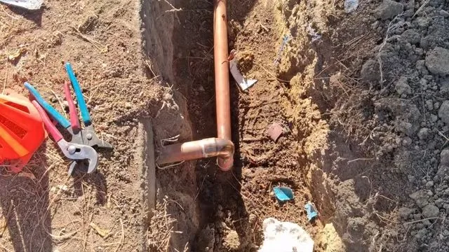 Copper pipe elbow in a yard trench beside pliers and a traffic cone
