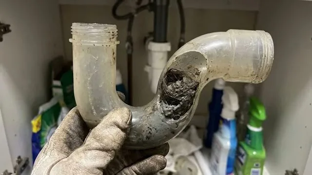 Gloved hand holding a plastic P-trap with a hole worn through the bend, in a cabinet with cleaning supplies