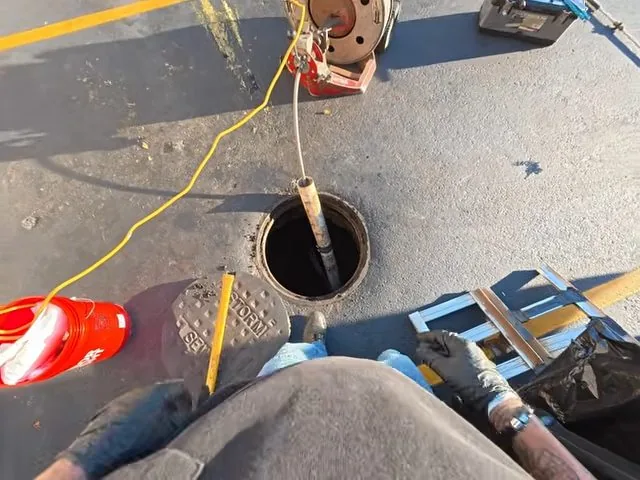 First-person view of a drain machine cable running down into a round sewer access opening in pavement