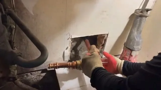 Gloved hands working on a copper pipe inside an opened section of drywall