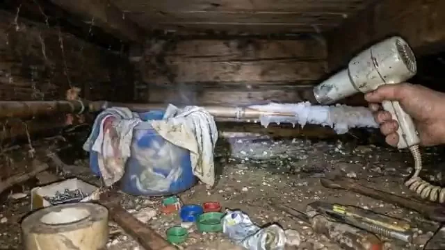 Hair dryer aimed at a frost-covered pipe in a dark crawlspace