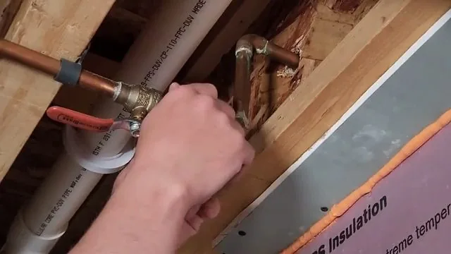 Hand turning a red-handled copper ball valve between floor joists