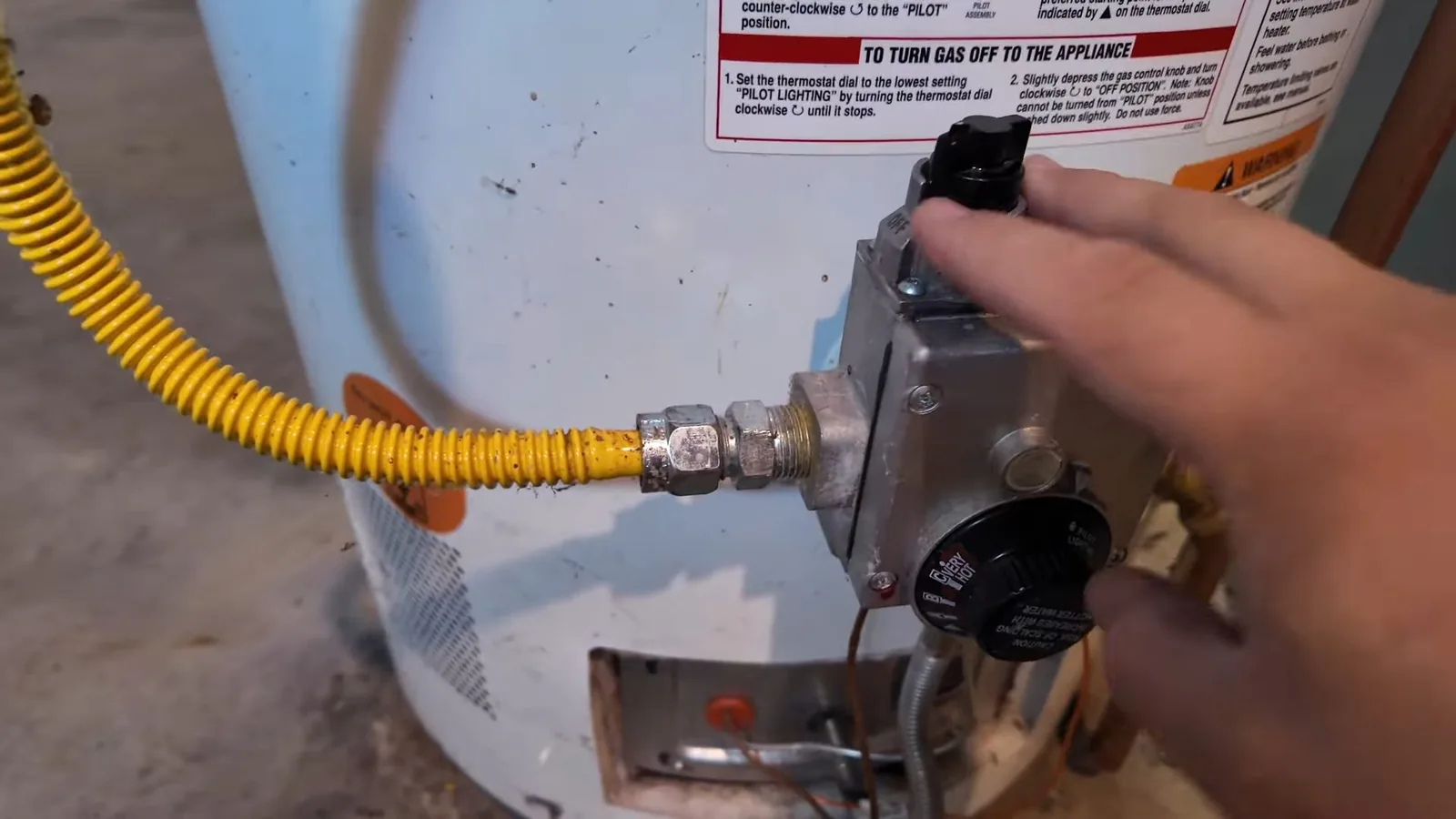 Hand on the gas control knob of a white water heater with a yellow flex line and an instruction label