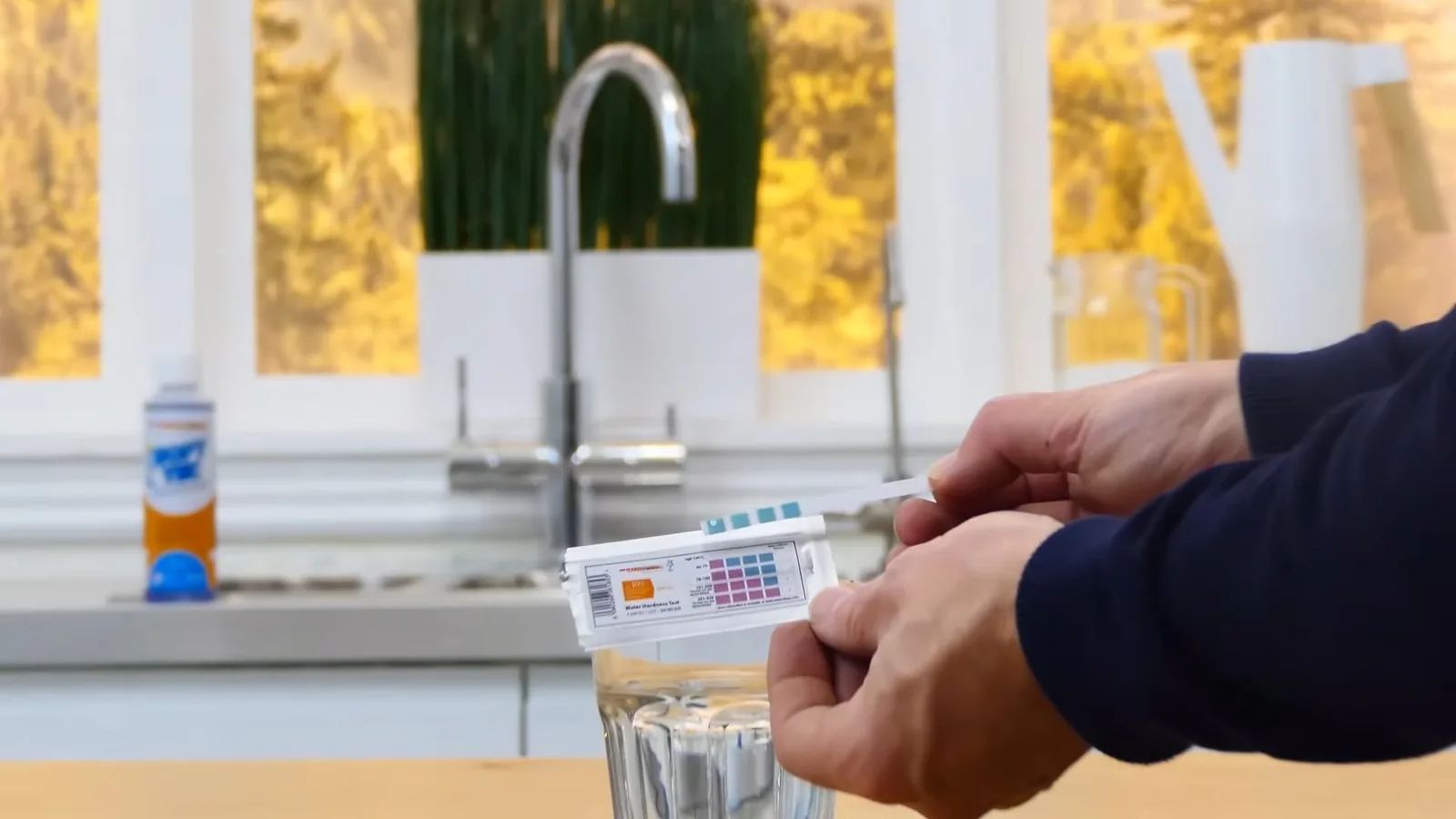 Hands holding a water hardness test strip over a glass at a kitchen tap