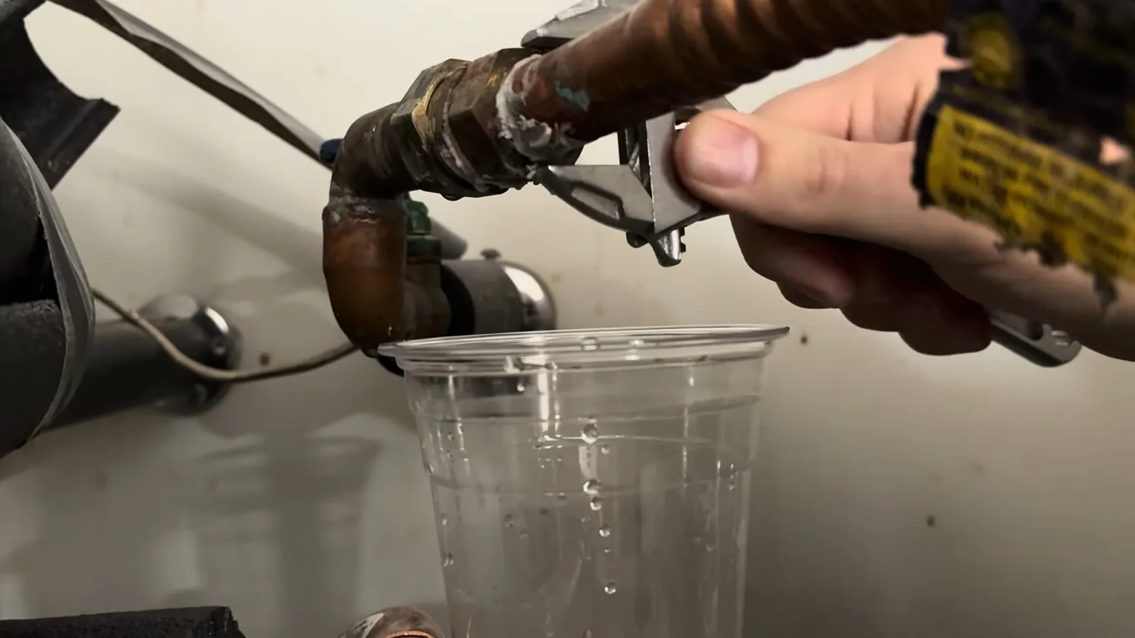 Hand with an adjustable wrench on a copper fitting at a heater with a clear cup held under it