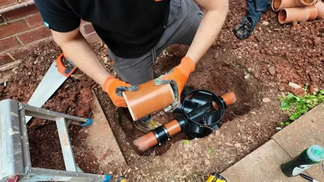 Man in a pit in a yard with orange sewer pipe parts, a saw and a ladder around him