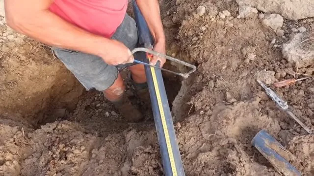 Man measuring a section of black pipe lying in an excavated hole