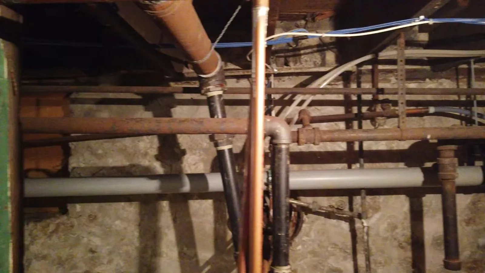 Overhead view of old cast iron and galvanized pipes running under a house in a dim crawlspace