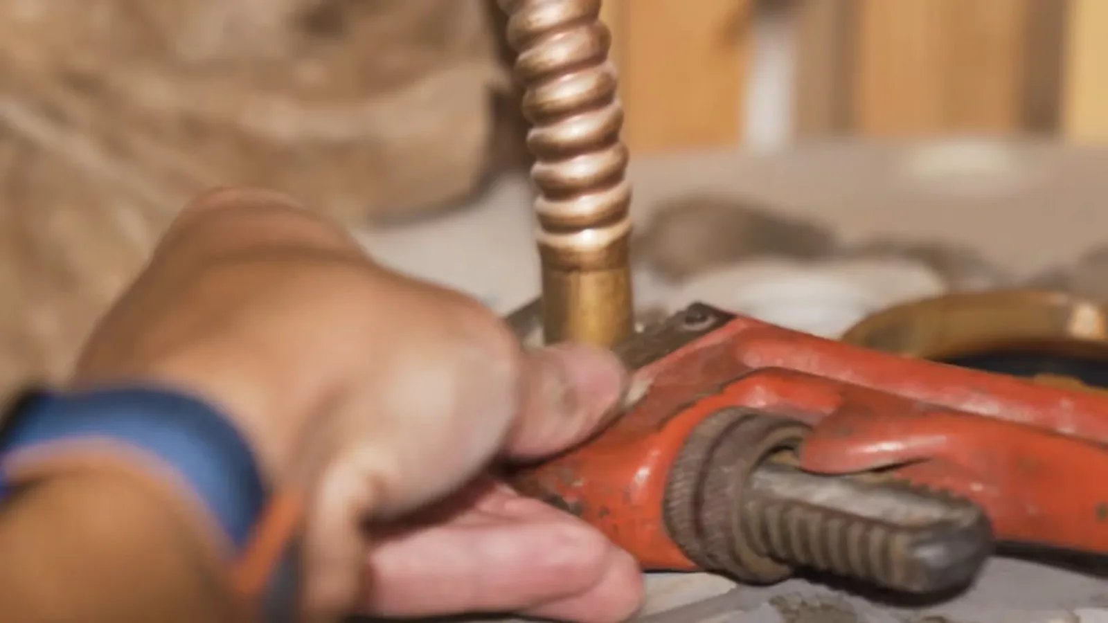 Hand with an orange pipe wrench on a corrugated copper water connector above a fitting