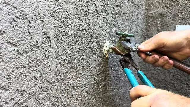 Hand with pliers and a wrench working on an outdoor hose bib on a stucco wall