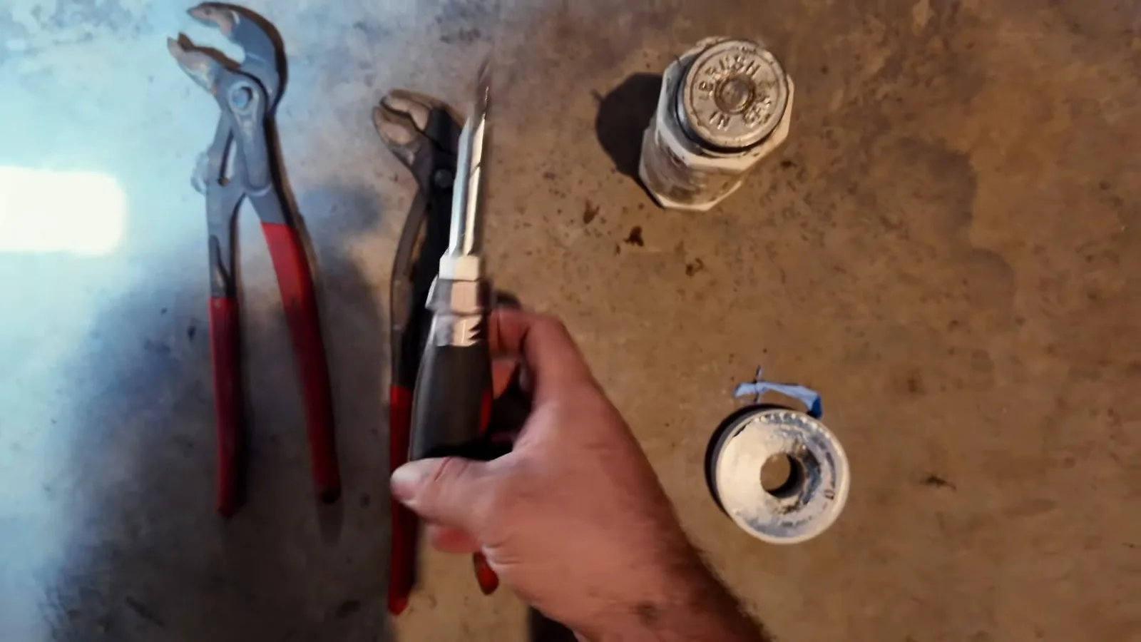 Channel-lock pliers, a roll of thread seal tape and threaded pipe fittings laid on the ground beside a hand