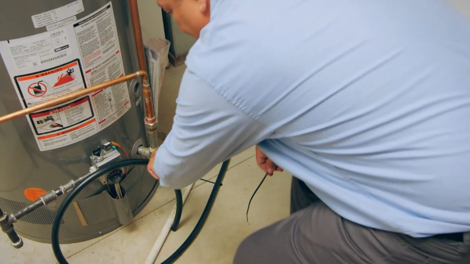 Man crouching at the base of a gas water heater with a drain hose attached