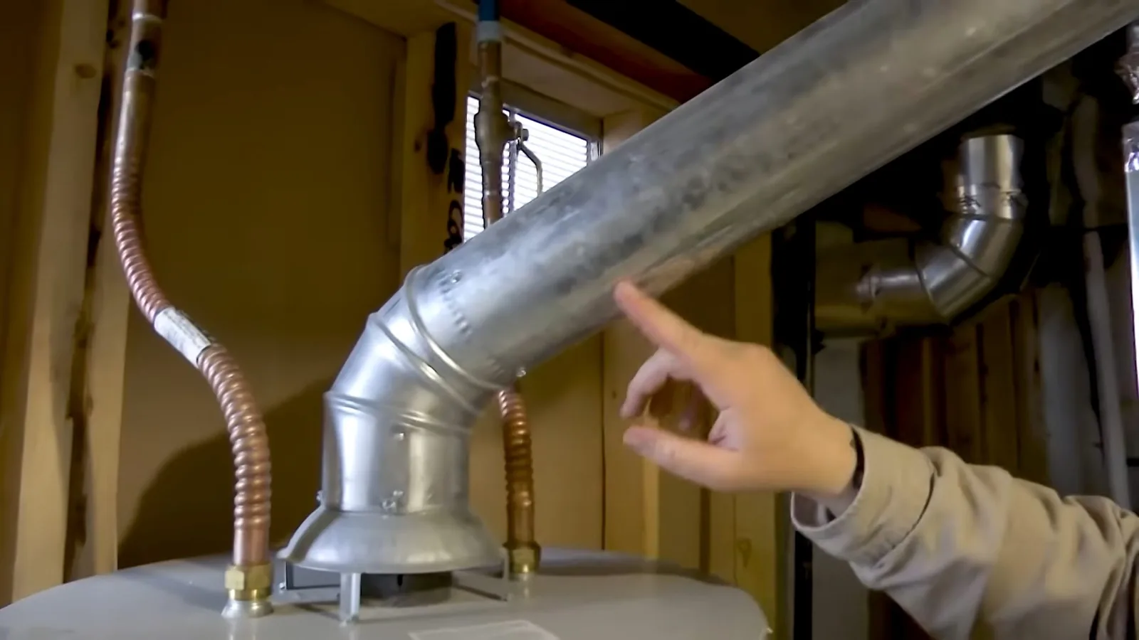Hand pointing at the vent pipe on top of a tank water heater with flexible gas lines nearby