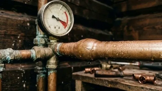 Round pressure gauge mounted on a copper water line with fittings and tools on a bench behind