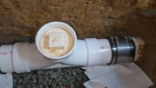 White PVC cleanout tee with a square-headed plug sitting in a trench on gravel