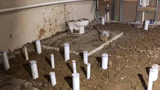 White PVC drain stubs standing in the dirt floor of a basement with a toilet in the background