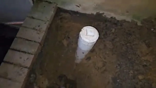 White PVC cleanout riser with a square plug standing in dug soil beside a brick wall