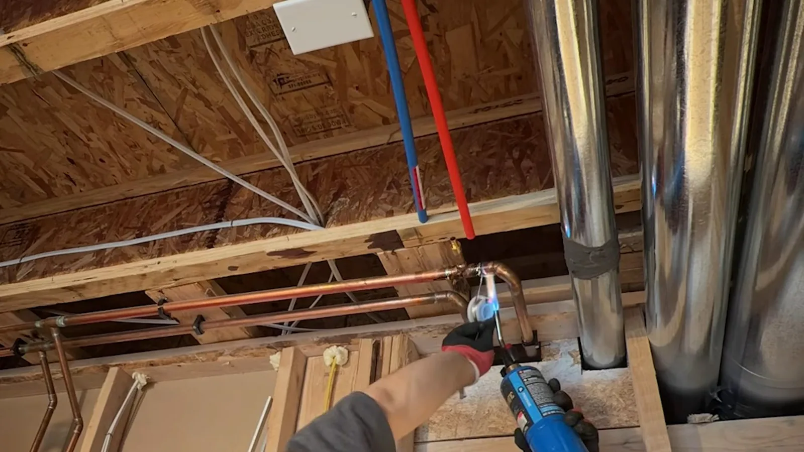 Red and blue PEX lines running through open floor joists beside copper pipe and a hand holding a torch