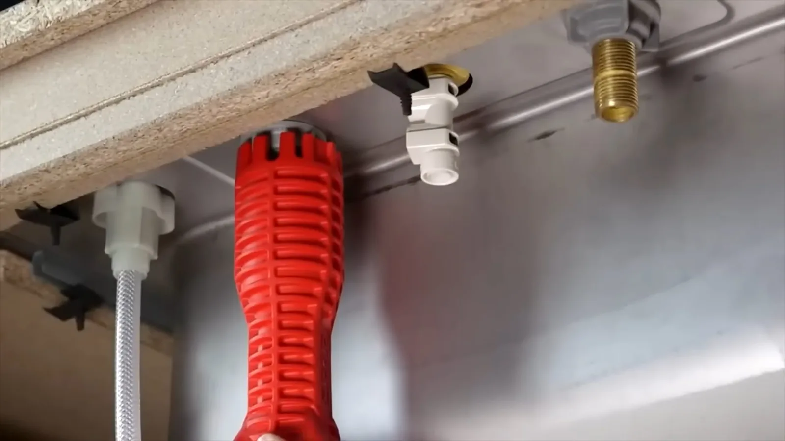 Red basin wrench resting under a kitchen sink beside supply connections