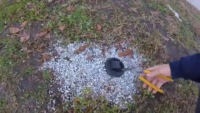 Hand with yellow-handled pliers beside an opened black cleanout set in gravel on a lawn