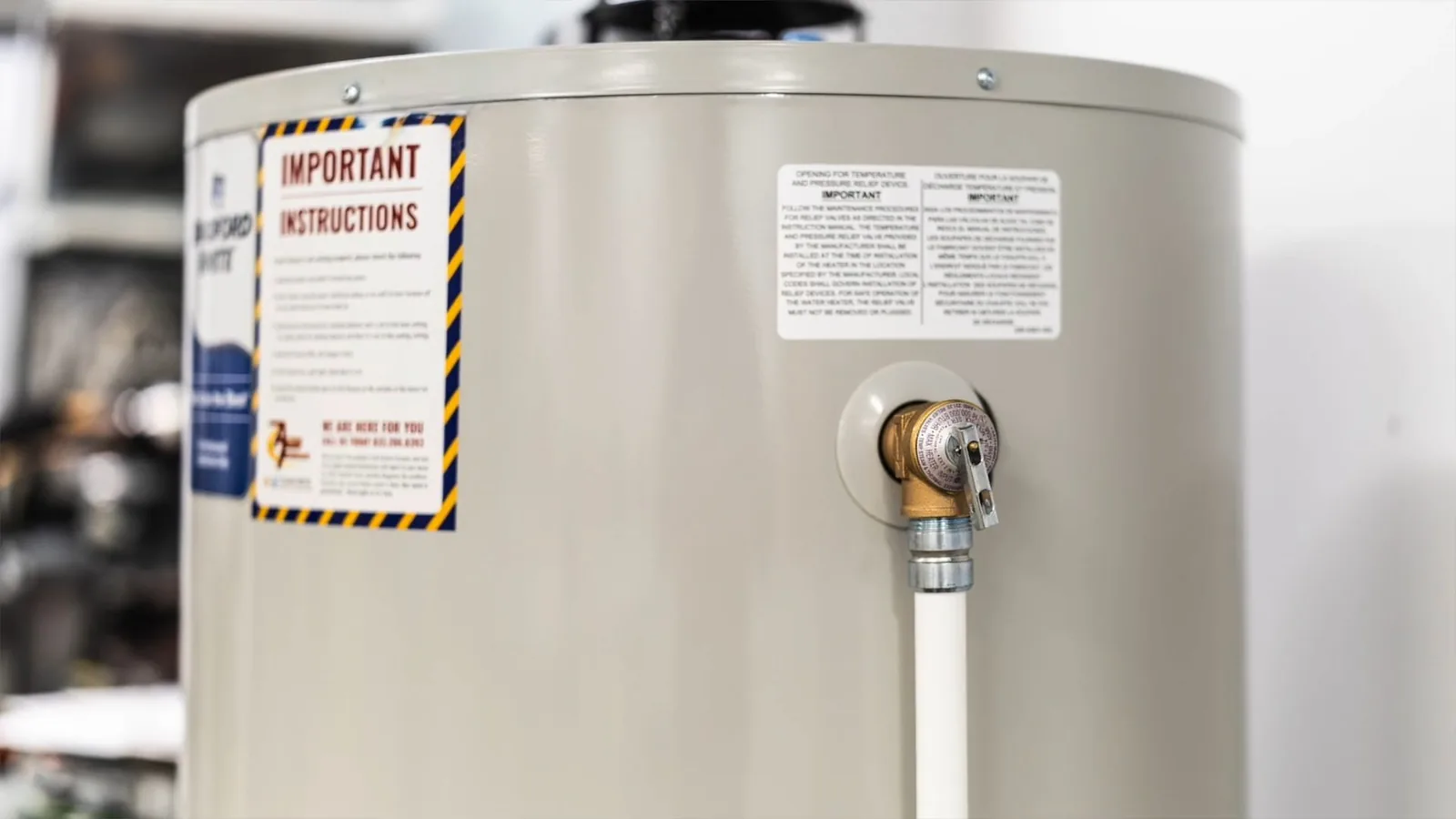 Upper part of a tank water heater with a yellow-and-black instruction label and a white supply line at the inlet