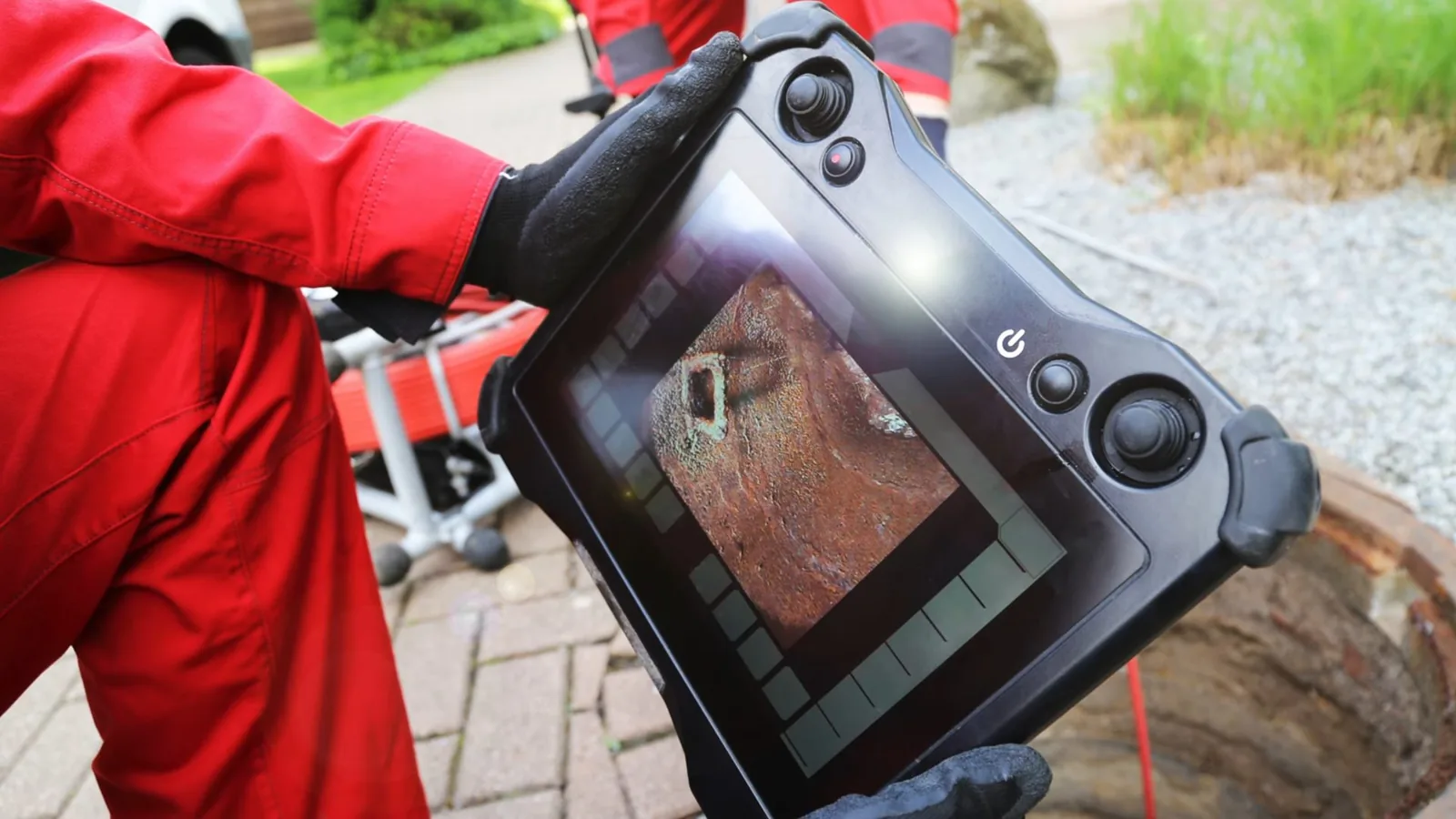 Technician in a red coverall holding a handheld camera monitor on a paved area beside a pipe opening