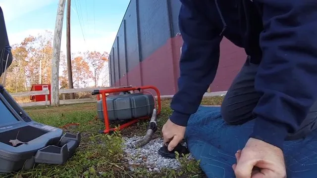 Technician kneeling at a gravel-ringed cleanout in a grassy yard beside a camera reel and monitor