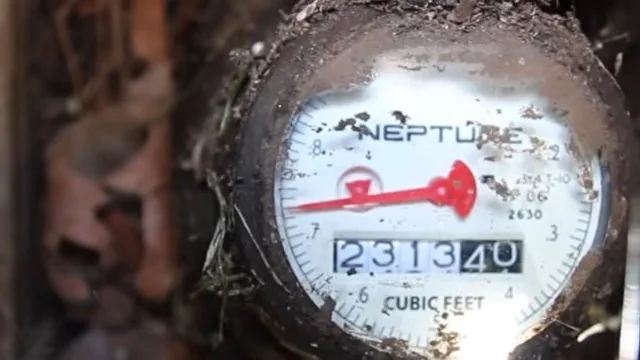 Close-up of a dirt-spattered water meter dial showing the red sweep hand and cubic-feet digits