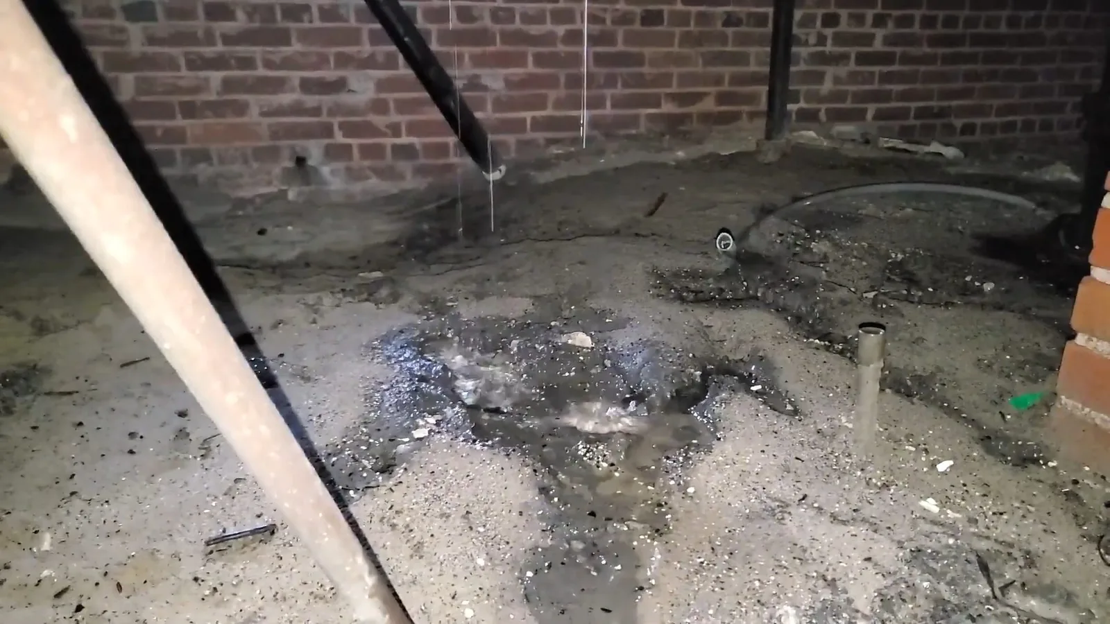 Wet, debris-covered ground in a crawlspace along a brick foundation wall with a short pipe stub