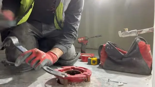 Gloved hands with a wrench at a red toilet flange fixed to a concrete floor