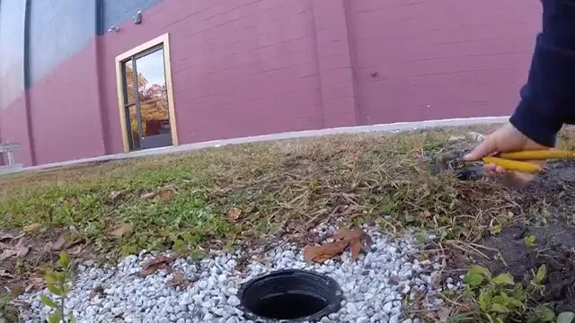 Cleanout opening in a gravel patch at the edge of a lawn with a hand holding pliers beside it